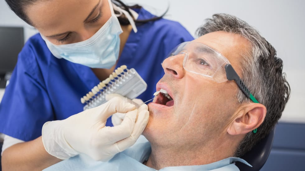 A female dental assistant in blue scrubs and a face mask holding a tooth shade guide up to a male patient's teeth to find a color match for a cosmetic procedure.