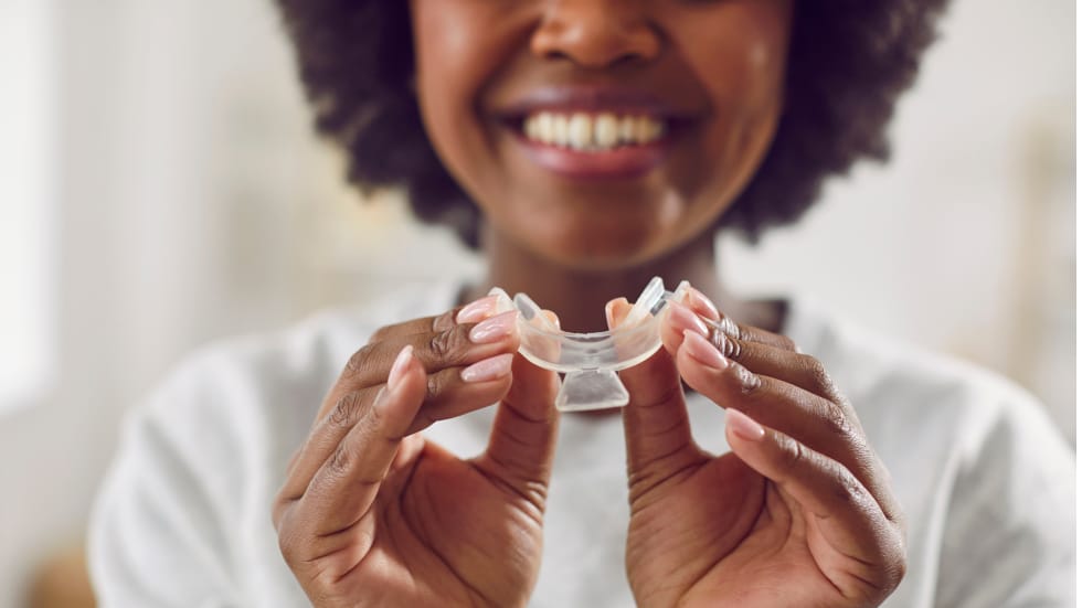 A close-up of a smiling woman holding a clear plastic dental mouth tray or mouthguard with both hands.